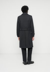 Manteau en laine gris au design ajusté, avec une taille ceinturée, un col haut et des poignets boutonnés, associé à un pantalon noir et des chaussures.