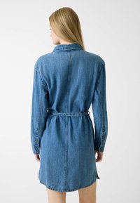 Abito camicia in denim color blu medio, con maniche lunghe, vita con cintura, polsini con bottoni e taglio dritto con orlo arrotondato.