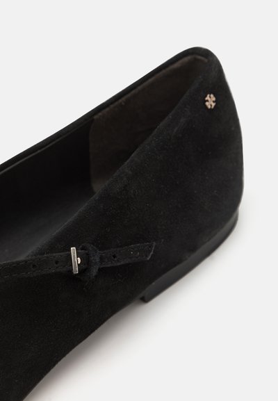 Chaussure plate en suede noir avec un bout rond, ornée d'un petit nœud et d'un détail en métal avec le logo sur le côté. Doublure intérieure lisse.