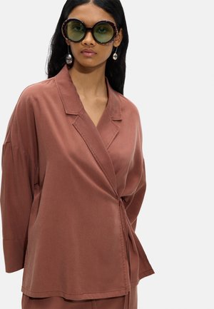 Kobieta nosząca duże okrągłe okulary przeciwsłoneczne, srebrne kolczyki i luźną, rdzawą kurtkę typu wrap na jednolitym tle.