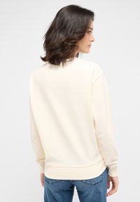 Felpa beige chiaro con una vestibilità comoda, polsini e orlo a costine; texture morbida; indossata con jeans blu, vista da dietro.