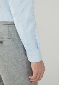 Camicia azzurro chiaro con polsini a bottone, caratterizzata da una texture liscia. Abbinata a pantaloni grigi con texture e tasca laterale.