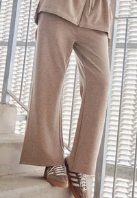 Beżowe szerokie spodnie dresowe z miękkiego materiału, z bocznymi rozcięciami. W zestawieniu z brązowo-białymi sneakersami na betonowych schodach.