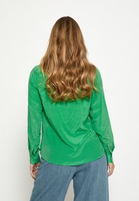 Grøn skjorte med lange ærmer i et glat stof med en buet kant, knapmanchetter og detaljer med bagplissering. Kombineret med blå bukser med vide ben.