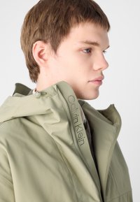 Olive green hooded jacket made of smooth, lightweight material. Features a zip closure and printed branding on the collar.