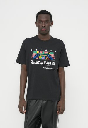 Camiseta negra de algodón con un gráfico colorido que incluye banderas y un diseño de fútbol, etiquetada como "adidas FIFA WorldCup USA 94." Mangas cortas, ajuste holgado.