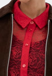 Red embroidered blouse with rose pattern, featuring a buttoned collar, surrounded by a brown suede jacket with silver zipper detail.