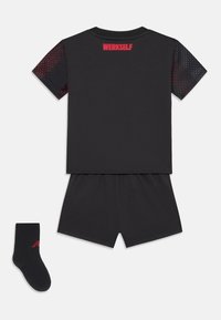 Conjunto atlético preto com uma camisa de mangas curtas com detalhes vermelhos e ombros em malha, combinado com calções a condizer e uma meia preta.