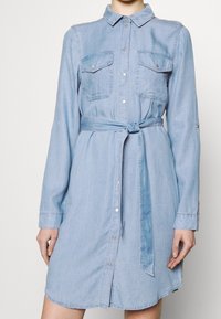 Ljusblå denimklänning med långa ärmar, knappstängning fram, två bröstfickor och ett matchande midjeband. Har en rundad nederkant.