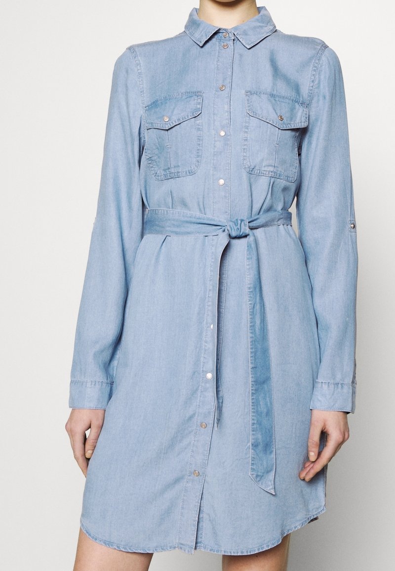 Ljusblå denimklänning med långa ärmar, knappstängning fram, två bröstfickor och ett matchande midjeband. Har en rundad nederkant.