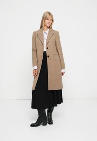 Cappotto in misto lana beige con rever a lancia, design monopetto e bottoni neri. Abbinato a una gonna nera plissettata e stivali neri con tacco.