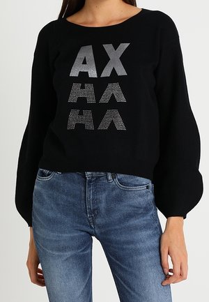 Frau trägt einen schwarzen Pullover mit Puffärmeln und silbernen Buchstaben "AX" sowie gemustertem "AA" vorne, kombiniert mit blauen Jeans.