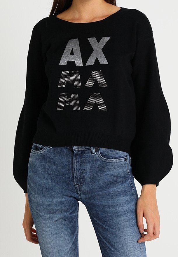 Femme portant un pull noir à manches bouffantes avec des lettres argentées "AX" et un motif "AA" à l'avant, assorti à un jean bleu.
