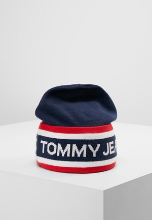 Granatowa czapka z prążkowanym paskiem w kolorach czerwonym, białym i niebieskim z napisem "TOMMY JEANS", złożona na białej powierzchni.
