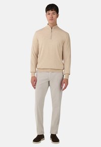 Pull en maille beige avec un col montant zippé, associé à un pantalon gris clair texturé et des chaussures à enfiler marron foncé.