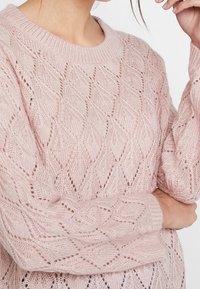 Maglione lavorato a maglia di colore rosa chiaro con un motivo a diamante in pizzo, scollatura rotonda e polsini a coste. La texture sembra morbida e a punto aperto.