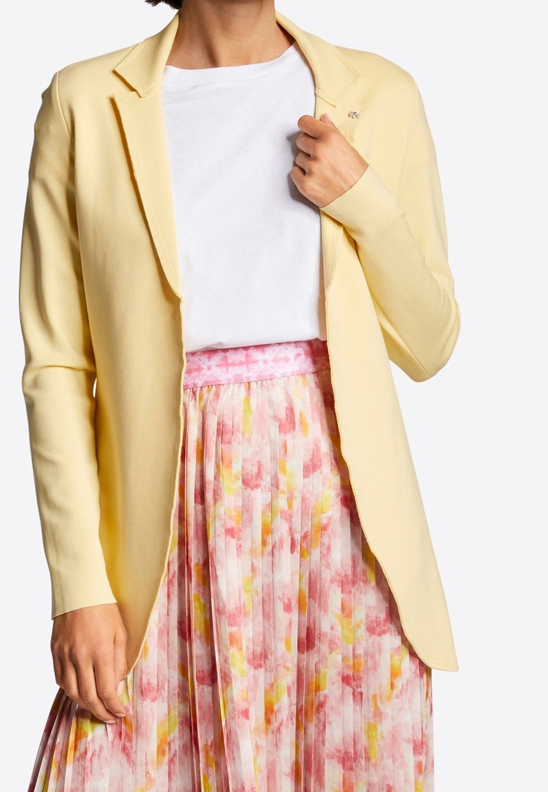 Personne portant un blazer jaune clair, un t-shirt blanc uni et une jupe plissée avec des motifs aquarelle rose, jaune et blanc.