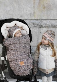 Blommönstrad babyfilt i en barnvagn, med gräddfärgad foder. Barn bredvid, iklädd en vit väst med dekorativ krage och matchande hatt.