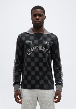 Homme portant une chemise à manches longues à carreaux noirs et gris de Champion F.C. avec col, et un pantalon de survêtement gris clair devant un fond uni.