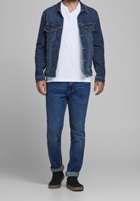 Denim jacket with button-up front, dark wash, and two chest pockets, worn over a white V-neck t-shirt, paired with rolled blue jeans and black shoes.