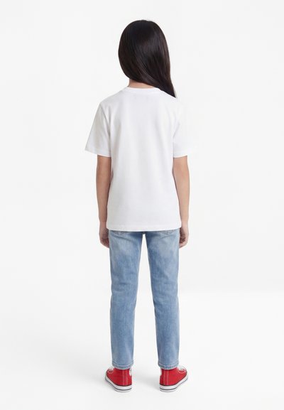 Enfant aux longs cheveux foncés portant un t-shirt blanc uni, un jean bleu clair et des baskets rouges, debout de dos sur un fond blanc.