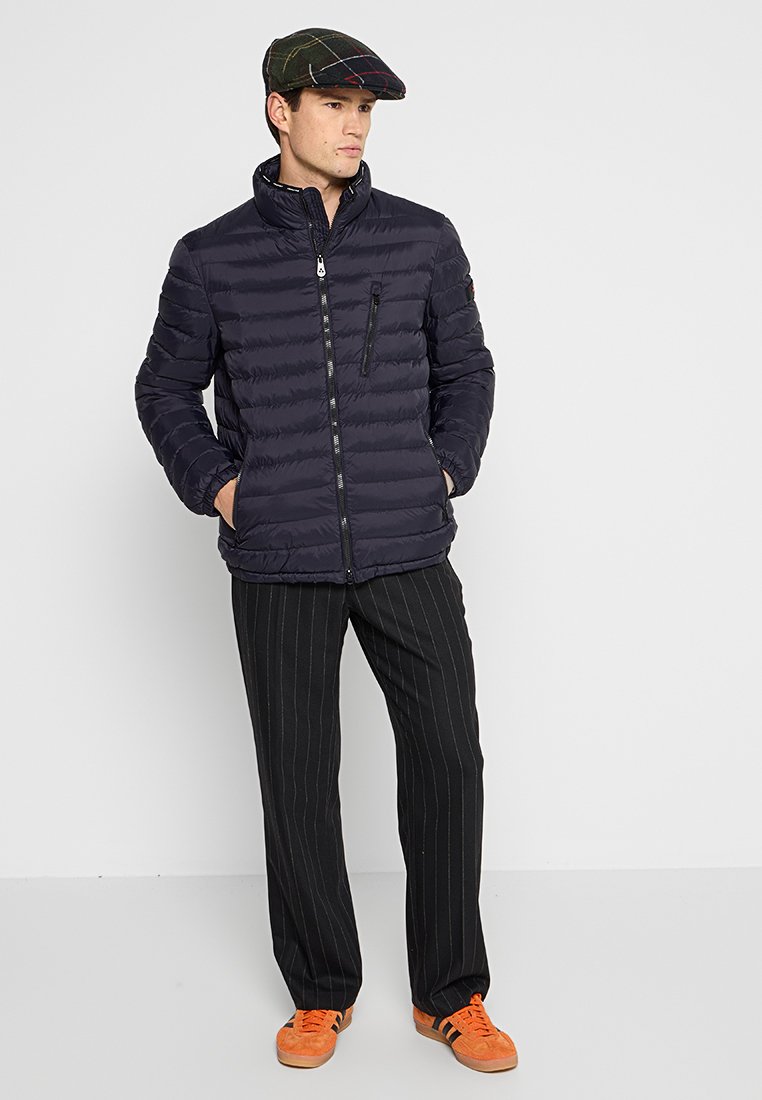 Man met een zwarte pufferjack, een geruite platte pet, gestreepte broek en feloranje sneakers, staand tegen een effen achtergrond.