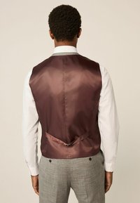 Burgundijas satīna veste ar gludu tekstūru, ar aizmugurējo jostu un pelēkiem rūtainiem biksēm. Balta krekla piedurknes ir redzamas.