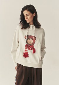 Svetlo béžová mikina s kapucňou s grafikou medvedíka, ktorý má červený pásikavý šál, mikina má šnúrkovú kapucňu a voľný strih.