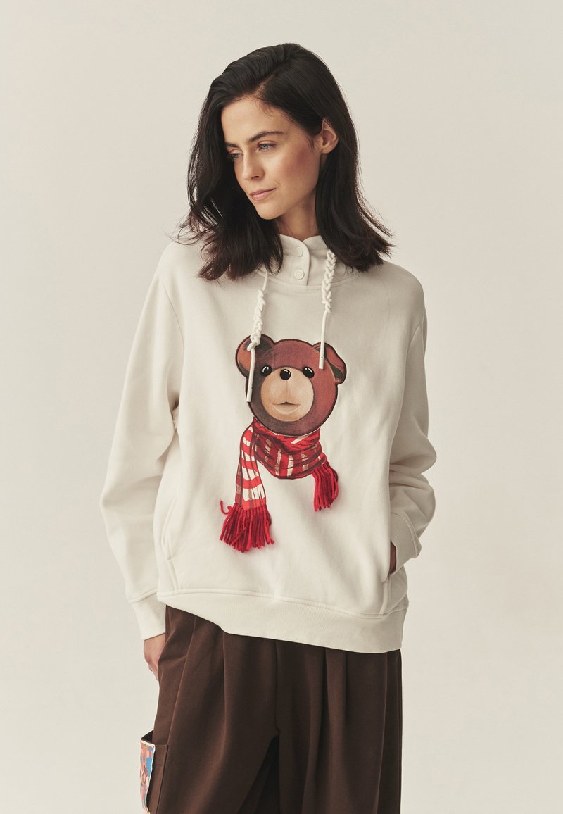 Svetlo béžová mikina s kapucňou s grafikou medvedíka, ktorý má červený pásikavý šál, mikina má šnúrkovú kapucňu a voľný strih.
