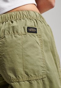 Olijfgroene cargobroek met een elastische tailleband, voorzien van een achterzak en een zwart gestikt merklabel. Glad, lichtgewicht stoffen textuur.