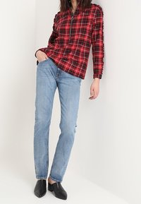 Rood en zwart geruit shirt met een halve rits en textuuraccenten op de schouders, gecombineerd met slanke blauwe denim jeans en zwarte instappers.