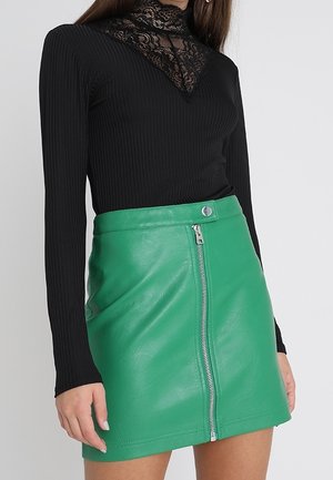 Femme portant un haut noir à manches longues avec détail en dentelle et une mini-jupe en cuir verte taille haute avec une fermeture éclair à l'avant.