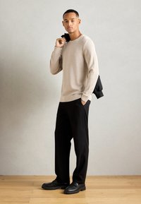 Pull en tricot beige clair, pantalon noir et chaussures noires avec une semelle épaisse. Le mannequin tient une veste noire sur son épaule. Texture lisse.