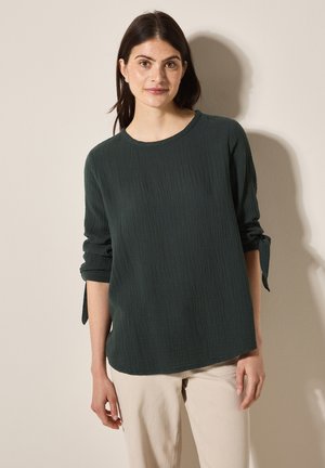 Chemise à manches longues texturée vert foncé avec un col rond et des détails de manches nouées. Portée avec un pantalon beige clair.