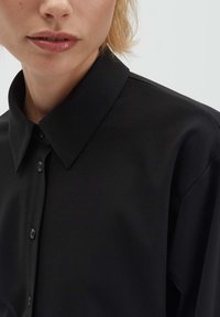 Camicia nera con bottoni, con collo classico, tessuto liscio e una leggera lucentezza. Include bottoni neri e vestibilità rilassata.