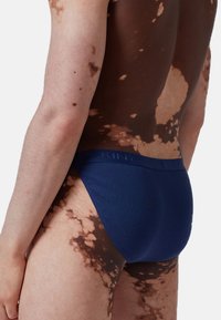 Slip da nuoto blu navy con un tessuto testurizzato e una vita elasticizzata con dettagli in rilievo del logo, mostrano un primo piano della visuale posteriore.