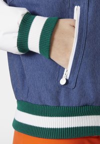 Denim jack met witte mouwen, groene en witte geribde manchetten. Heeft een ritszak en een gestructureerde stof. Oranje detail aan de onderkant.