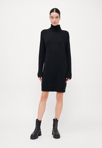 Melna adīta kleita ar augstu turtleneck un garām piedurknēm, kurai ir brīvs piegriezums un ribota tekstūra, apvārdota ar melniem potīšu zābakiem.