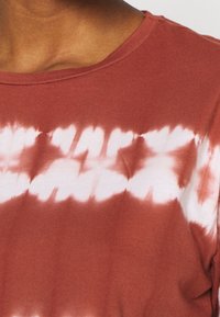 T-shirt en coton avec une base de couleur rouille, présentant un motif tie-dye blanc. L'encolure est ronde, avec une coupe décontractée et une texture douce.