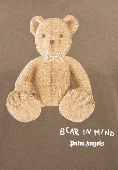 Γραφικό μπλουζάκι με μεγάλη, υφασμάτινη εικονογράφηση αρκούδου σε ανοιχτό καφέ, με λευκό γιακά και κείμενο από κάτω σε έντονη λευκή γραμματοσειρά.