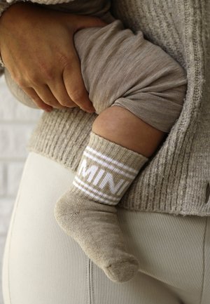 Adulte tenant un bébé vêtu d'un pantalon beige et de chaussettes beiges avec des rayures blanches et un texte partiel sur le revers.