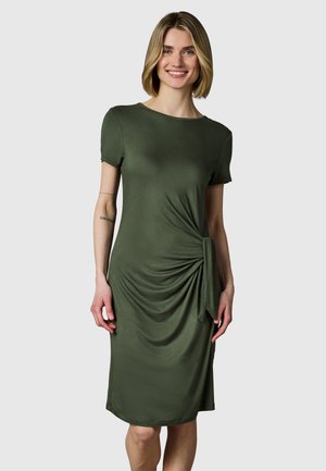 Lachende vrouw draagt een olijfgroene korte mouw knielange jurk met knoopdetail aan de zijkant, staand tegen een effen lichtgrijze achtergrond.