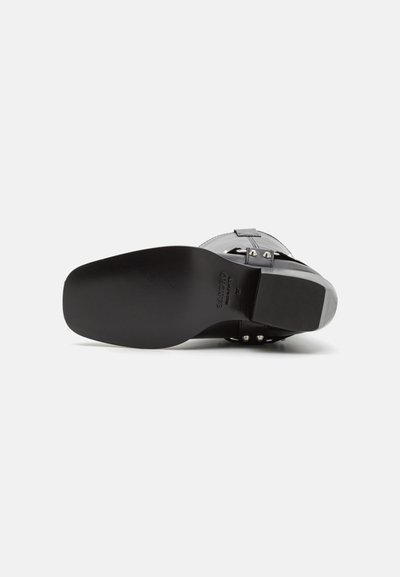 Scarpa in pelle nera con una texture liscia, suola piatta e tacco solido. I dettagli includono borchie metalliche sulla fascetta e una fibbia decorativa.
