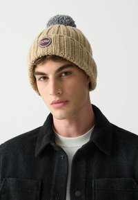 Beanie beige lavorato a maglia con pom-pom grigio e patch rotondo con logo Colmar. Design a coste testurizzate; tessuto morbido, vestibilità aderente.