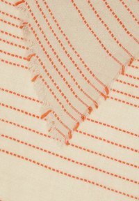 Textile tissé beige avec des rayures orange et des lignes en pointillés, présentant des bords frangés. Le tissu a une texture douce et un design décontracté.