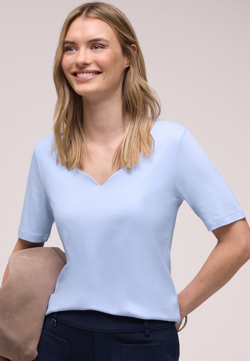 Mujer sonriente con cabello rubio hasta los hombros, vistiendo una camiseta de manga corta de cuello en V azul claro y pantalones oscuros, sosteniendo una chaqueta beige.