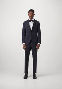 Uomo in piedi con un tuxedo blu scuro con rever nero, camicia bianca, papillon nero e scarpe eleganti nere contro uno sfondo semplice.