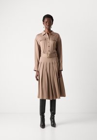 Beige mekko, jossa on pitkät hihat, napit edessä ja tuotettu hame, varustettu vastaavalla vyöllä, jossa on kultaiset yksityiskohdat. Päällä mustat polven yli ulottuvat saappaat.