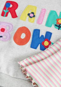 Grauer Sweatshirt mit mehrfarbig gestickten Buchstaben, die "RAINBOW" spelln, mit floralen Akzenten. Unter einer gestreiften pastellfarbenen Stofflage.