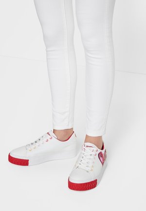 Białe trampki z czerwonymi teksturowanymi nakładkami na palce, czerwonym wnętrzem i różowymi wzorami w kształcie serca, noszone z białymi dopasowanymi spodniami na jednolitym białym tle.